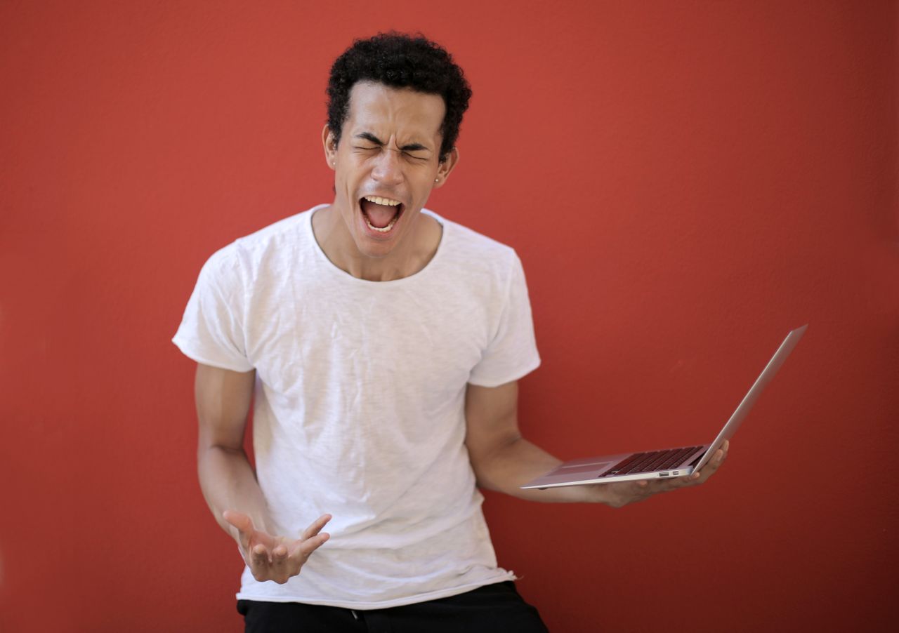 Guy with opened mouth and closed eyes screaming madly while standing with laptop against red background