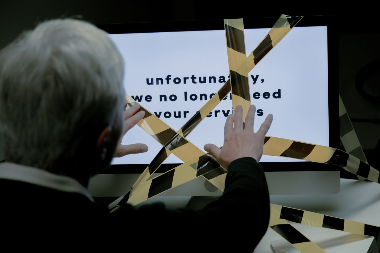 An elderly man reacts to a dismissal message on a computer, conveying ageism and unemployment themes.